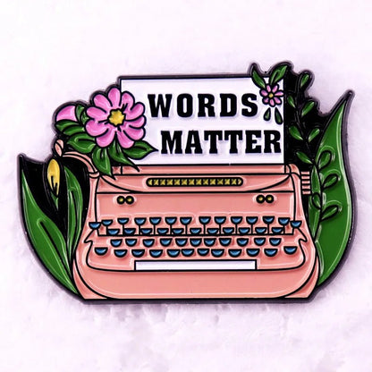 A charming enamel pin — a pink typewriter with floral accents and a paper reading WORDS MATTER in bold, perfect for Romance Readers or book lover badge collectors.