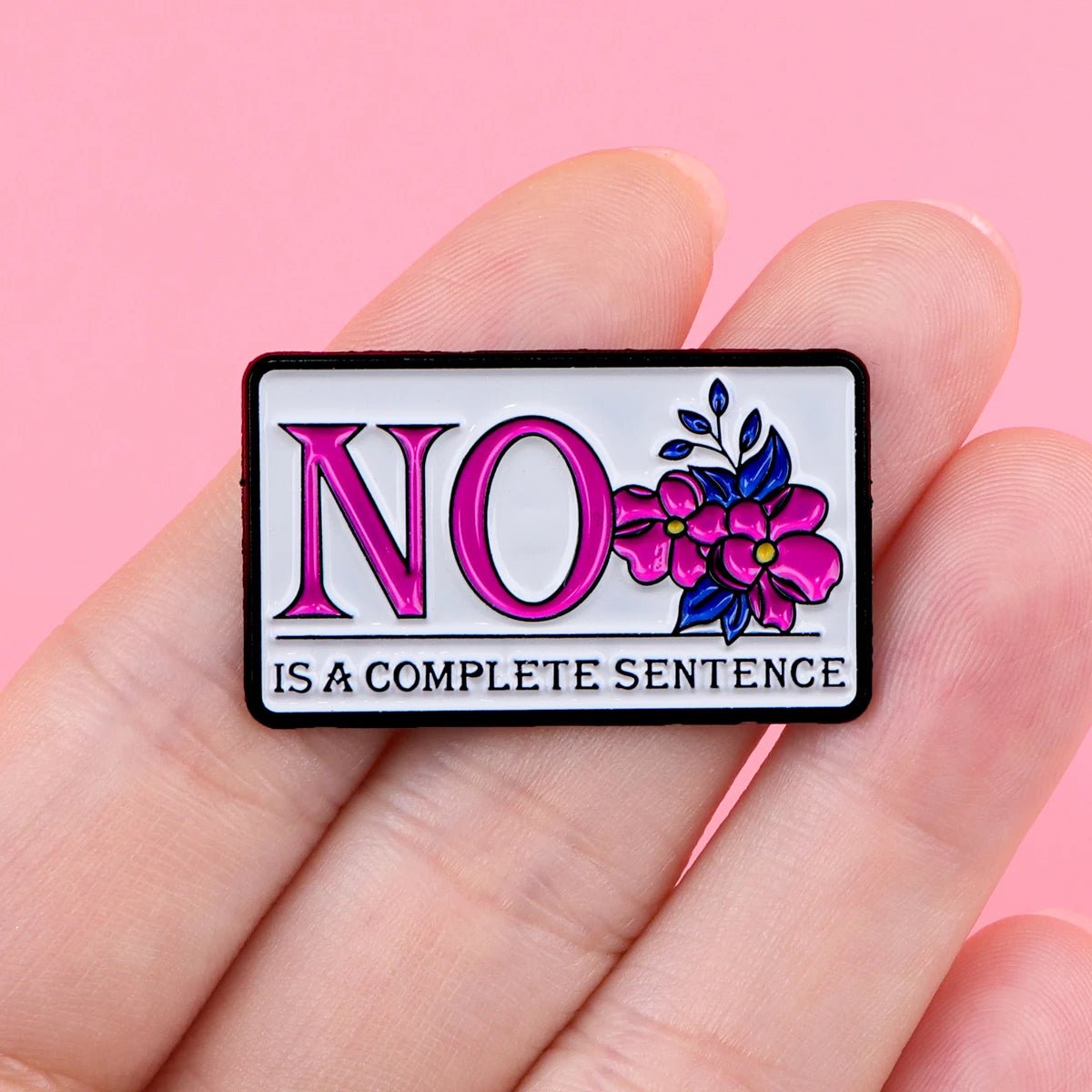 A hand holds a My Cutesy Era floral enamel pin reading NO IS A COMPLETE SENTENCE in pink and black letters, accented with pink flowers, set against a pink background—ideal for lovers of bold feminist accessories.