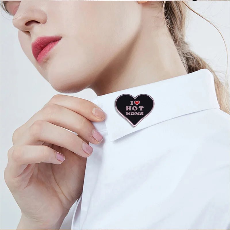 A person in a white collared shirt holds the collar, showcasing a I Love Hot Moms enamel pin shaped like a black heart with white and pink lettering.