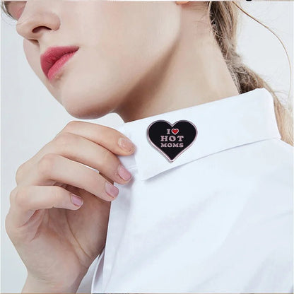 A person in a white collared shirt holds the collar, showcasing a I Love Hot Moms enamel pin shaped like a black heart with white and pink lettering.