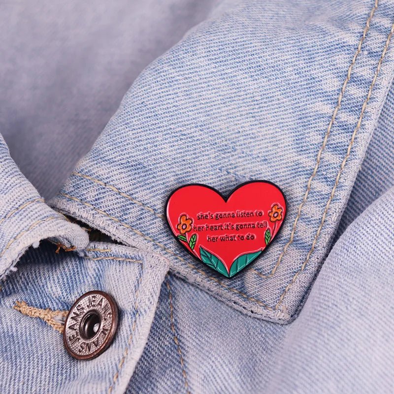 An enamel pin with a red heart, yellow flowers, and green leaves says shes gonna listen to her heart; its gonna tell her what to do—a perfect accessory for any denim jacket.n