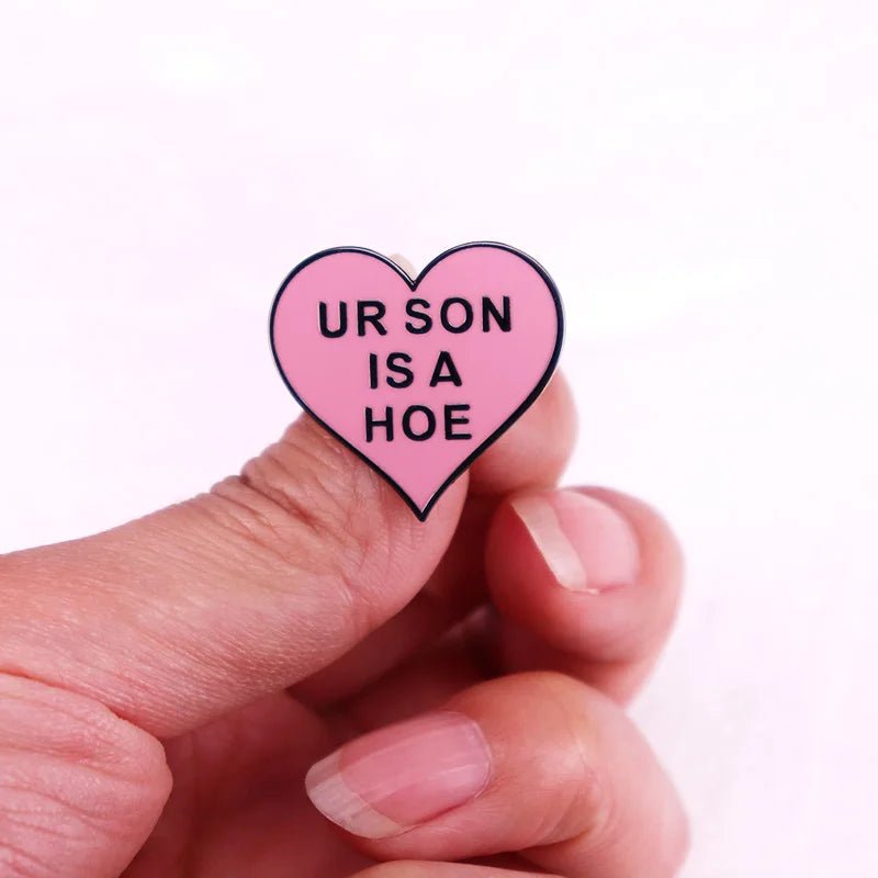 A hand holds a pink, heart-shaped enamel pin with black text reading UR SON IS A HOE against a plain light background.