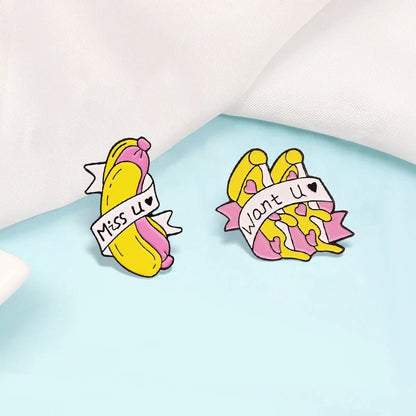 Two colorful enamel pins shaped like peeled bananas with banners—one says Miss U, the other Want U. These cute pink and yellow pins are displayed on a light blue background with a white cloth.