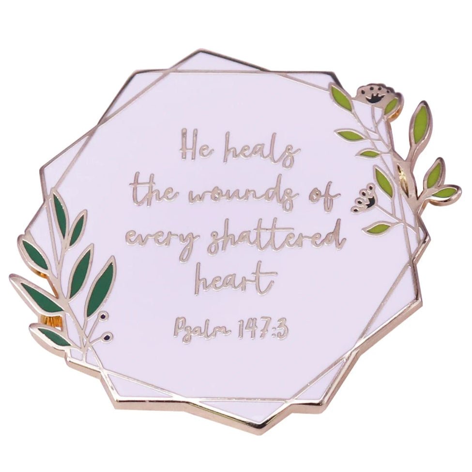 Enamel pin features green leaves, gold accents, and the comforting words: He heals the wounds of every shattered heart. Psalm 147:3.