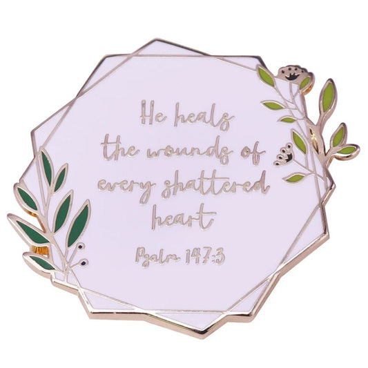 Enamel pin features green leaves, gold accents, and the comforting words: He heals the wounds of every shattered heart. Psalm 147:3.