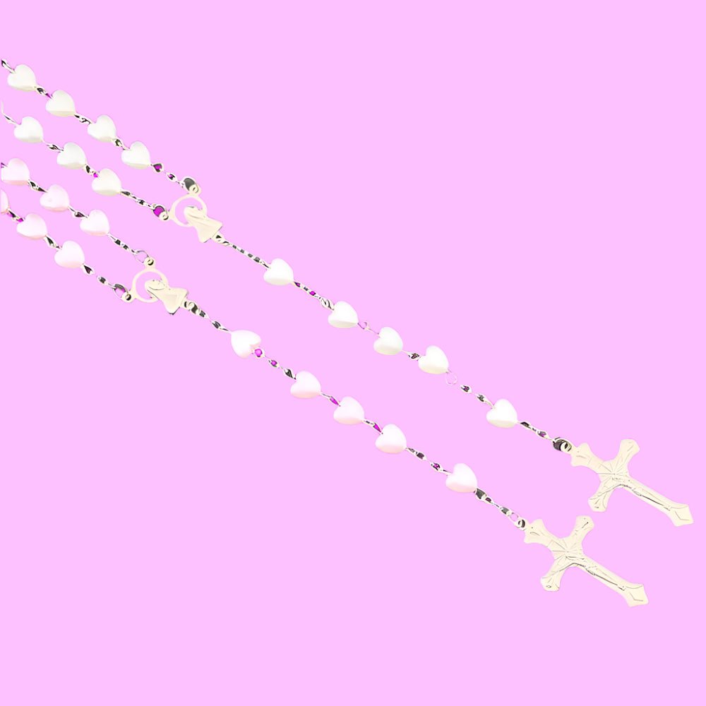 Two My Cutesy Era rosary necklaces with white heart-shaped beads and gold crucifixes, inspired by the Virgin Mary, are arranged diagonally on a pastel pink background.