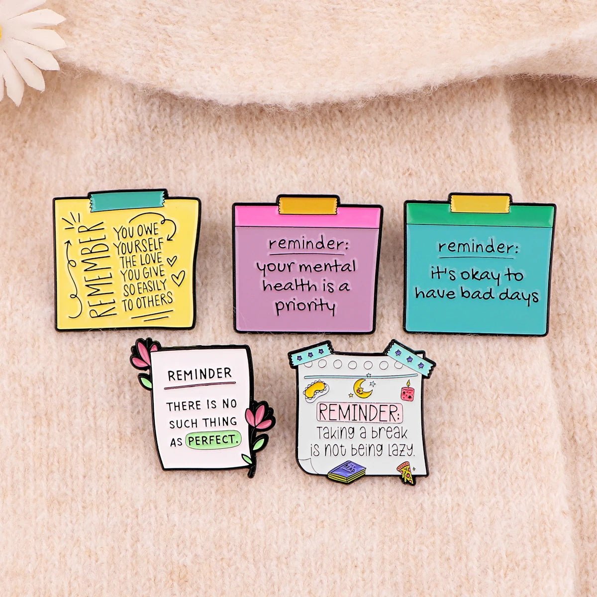 Five colorful enamel pins each with a unique motivational phrase, rest on soft beige fabric. These pins promote self-empowerment and mental health awareness through positive affirmations.