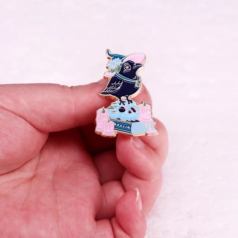 A hand holds a Gothic Book Crow enamel pin, featuring a black bird in a witch hat with pink candles, blue books, and a blue skull. Pastel colors and a magical theme make this unique accessory stand out.