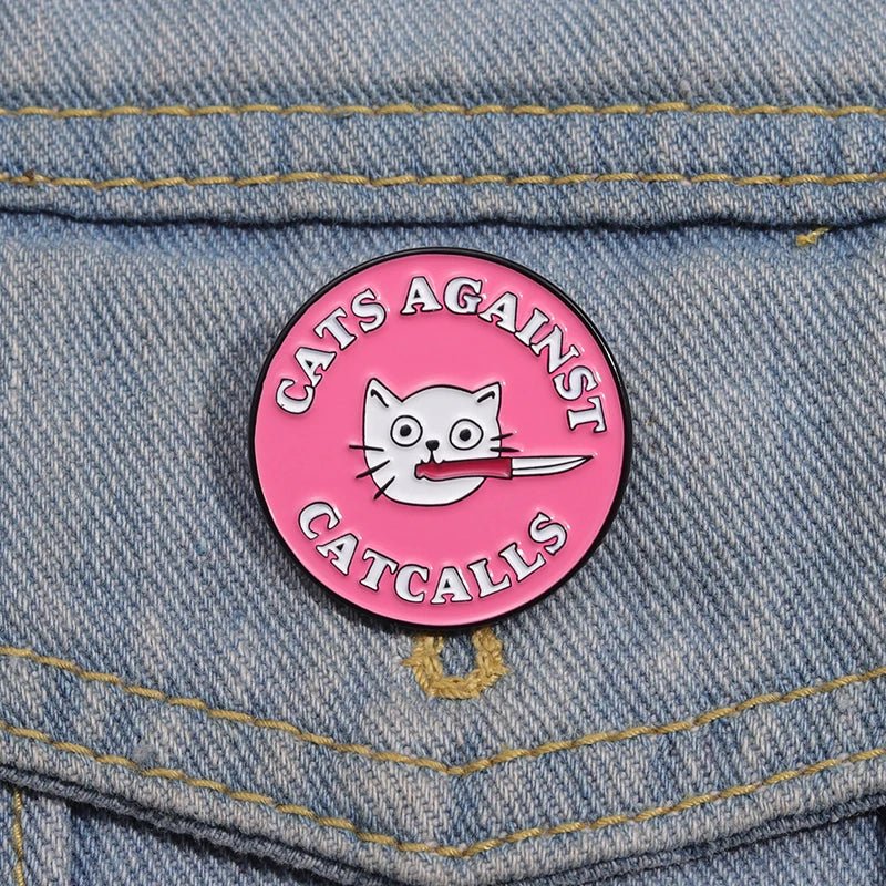 Pink enamel pin features a cartoon cat face with a knife in its mouth and Cats Against Catcalls text—a perfect feminist badge to show your stance on any blue denim fabric.