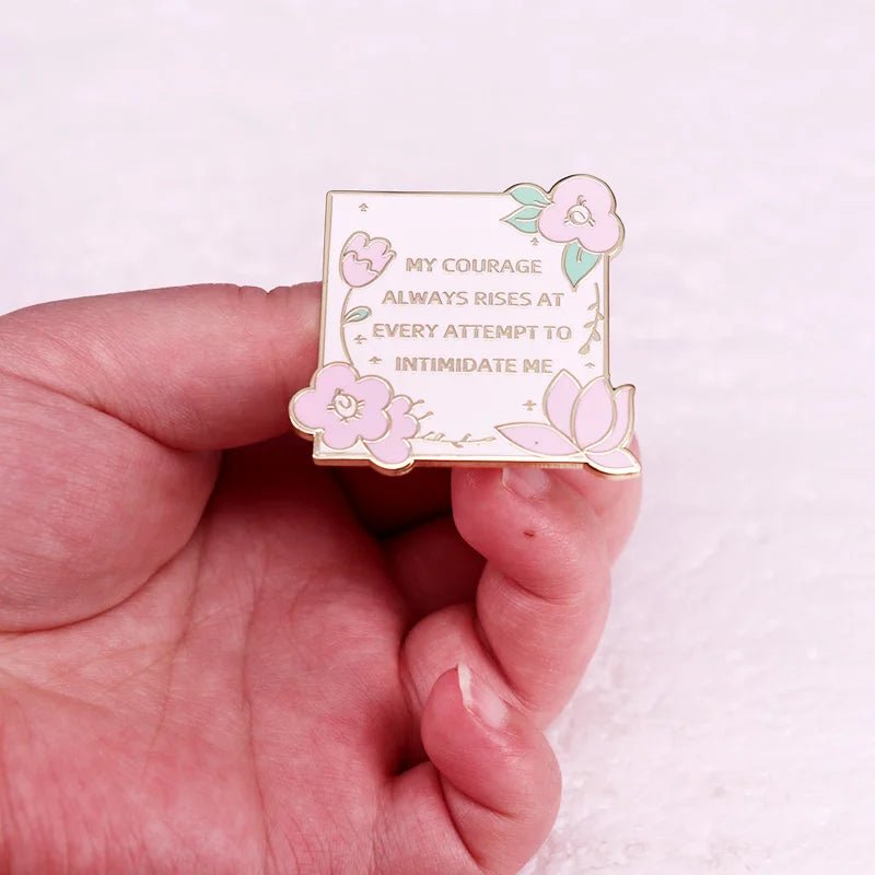 A hand holds a Kiss Me Later enamel pin adorned with pink flowers and leaves, featuring the quote: My courage always rises at every attempt to intimidate me.