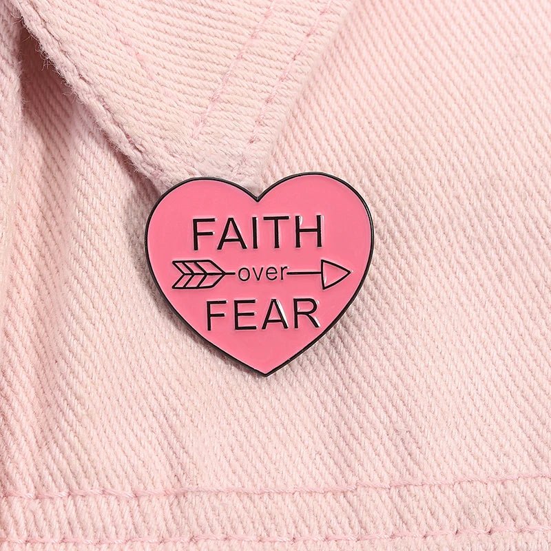 Pink heart-shaped enamel pin with Faith Over Fear and an arrow through over sits on light pink fabric.