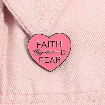 Pink heart-shaped enamel pin with Faith Over Fear and an arrow through over sits on light pink fabric.