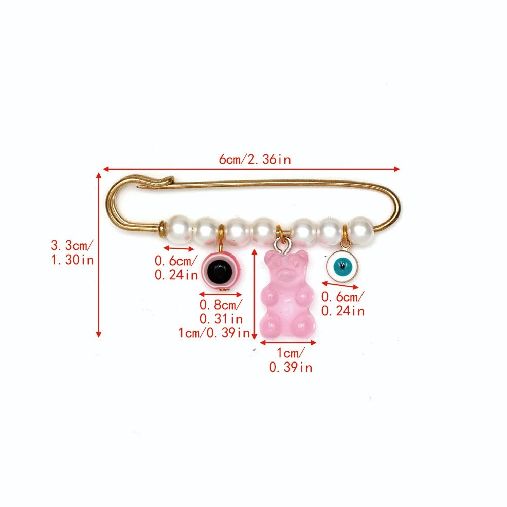 The Kiss Me Later gold safety pin is 6cm (2.4in) long and features five white beads plus three dangling charms—a 1cm (0.4in) pink bear and two evil eye charms—making it a perfect baby shower gift or unique protective pin.