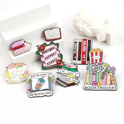 Enamel pins feature colorful book-themed designs perfect for romance readers—stacks of books, bookshelves, coffee, popcorn, flowers, and phrases like “Booktrovert” and “I want my coffee icy and my books spicy.”.