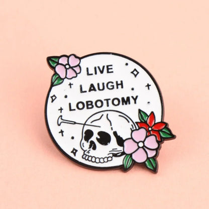 The Gay Skate round enamel pin features a skull with a nail in its head, surrounded by pink and red flowers. LIVE LAUGH LOBOTOMY stands out in bold on a white background—quirky flair for your collection!.
