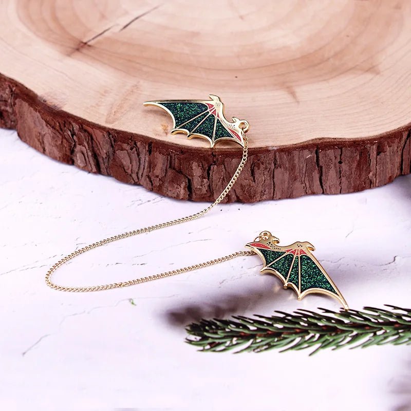 Gold chain earrings feature two glittery, green bat-shaped charms—resembling Green Dragon Wings—that sparkle and connect with a delicate chain, displayed on a light surface near a tree slice and pine branch.