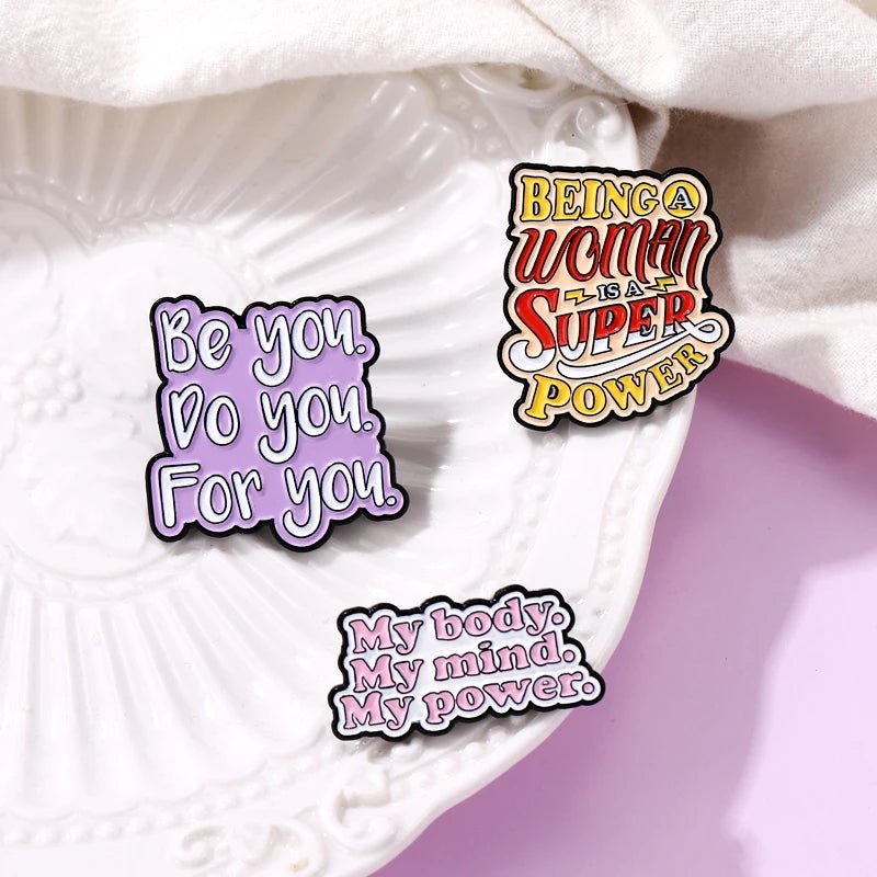 Three  enamel pins on a white plate feature self-love messages in bold, colorful fonts: a heart design, “Be you. Do you. For you.”, and “Being a woman is a super power.”.