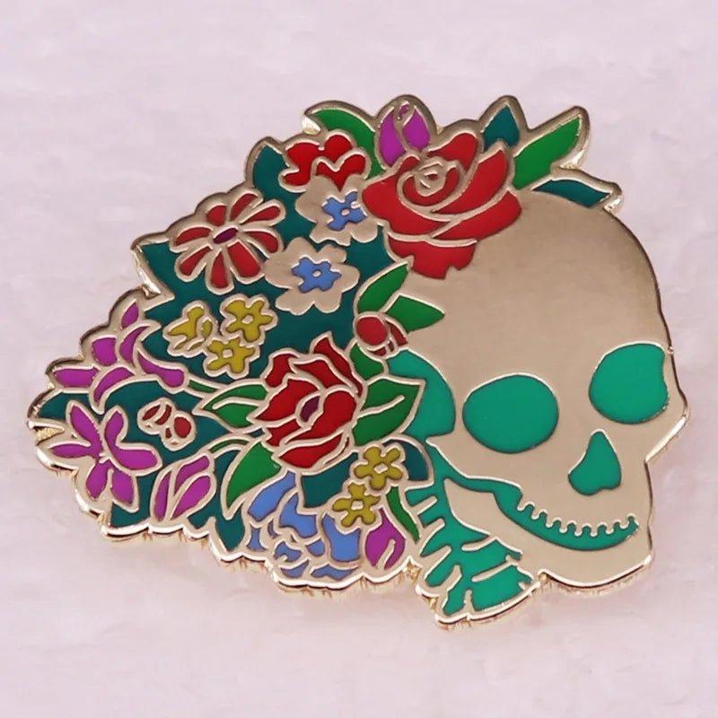 Enamel pin features a gold-outlined skull on the right and colorful flowers with red roses on the left, all set against a light background.