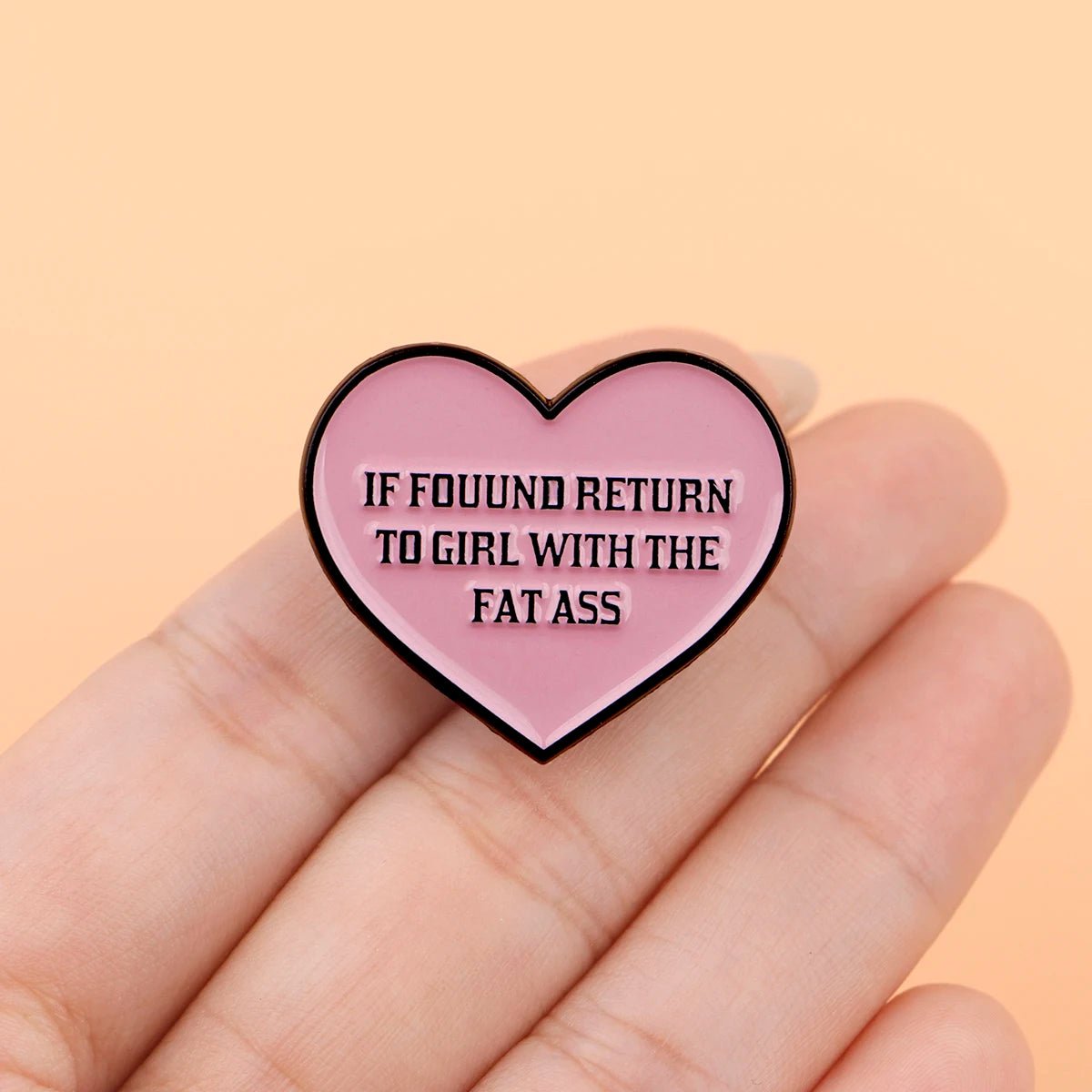 A hand displays a Kiss Me Later pink heart enamel pin reading IF FOUND RETURN TO GIRL WITH THE FAT ASS in bold black letters. The soft peach background highlights this novelty accessory.