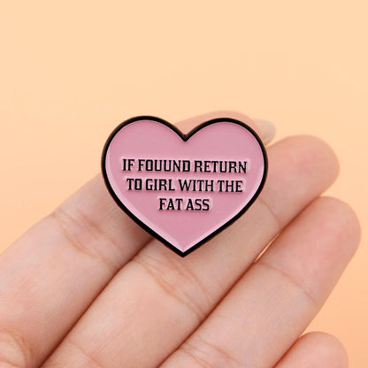 A hand displays a Kiss Me Later pink heart enamel pin reading IF FOUND RETURN TO GIRL WITH THE FAT ASS in bold black letters. The soft peach background highlights this novelty accessory.