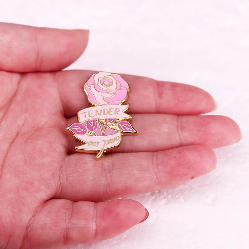 A hand holds a pink rose-shaped enamel pin featuring a gold finish and a “TENDER NOT TIMID” banner, standing out against a light textured background.