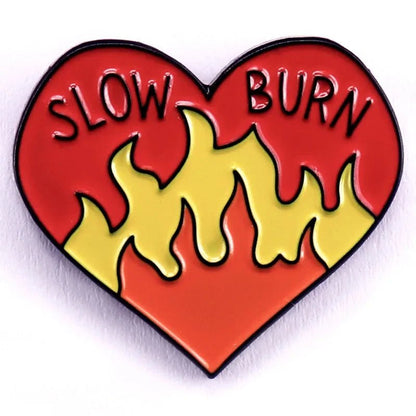 Red yellow and orange heart enamel pin features SLOW BURN at the top and bold yellow flames in the center, set against a vibrant red background.