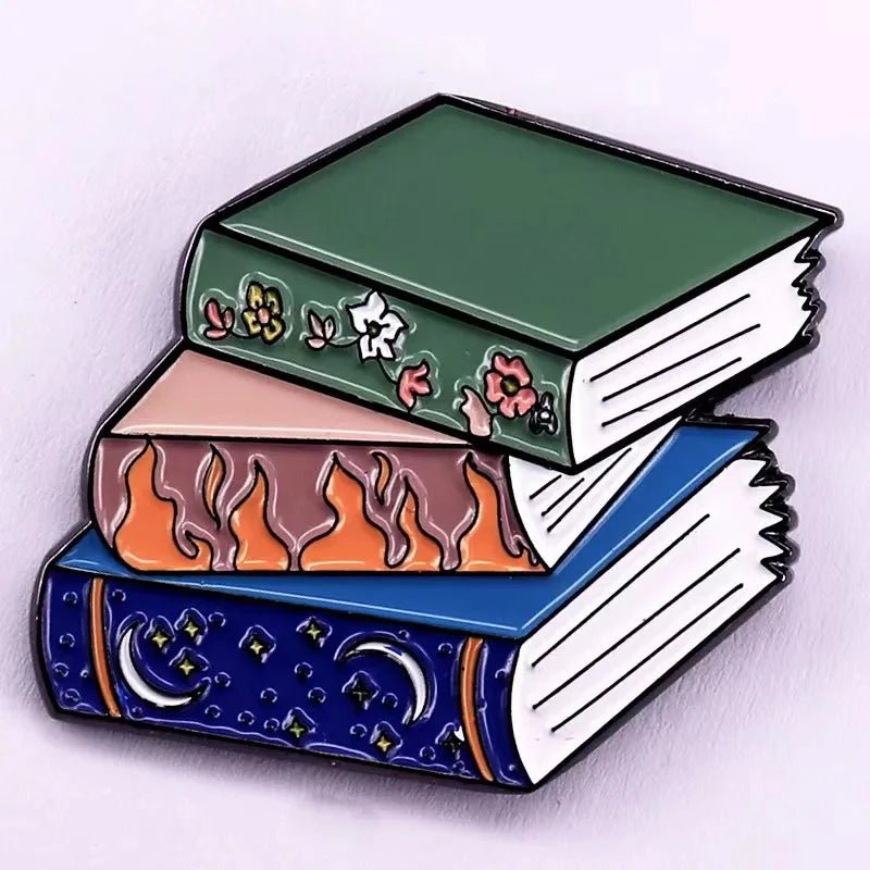 An illustration of three stacked books  a green floral book, a peach Romance Reader with flames, and a blue book with stars—perfect inspiration for enamel pins or badges for book lovers.