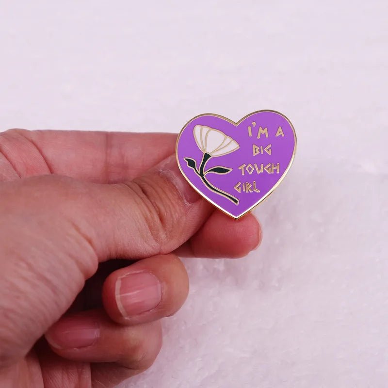 A hand holds a purple heart enamel pin with a white flower and yellow IM A BIG TOUGH GIRL text against a soft pink background—perfect for fans of unique, empowering pins.