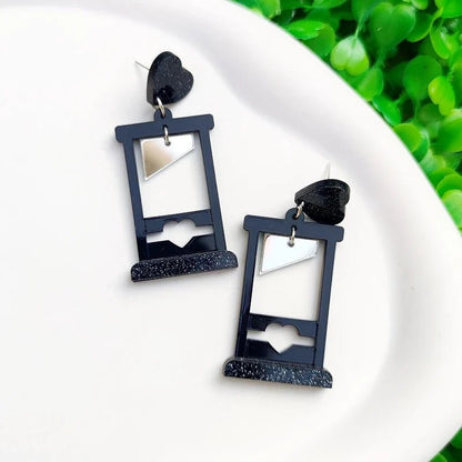 Gay Skate guillotine acrylic earrings feature black guillotines with heart cutouts, silver triangular blades, and black heart studs, displayed on a white surface with green leaves in the background.