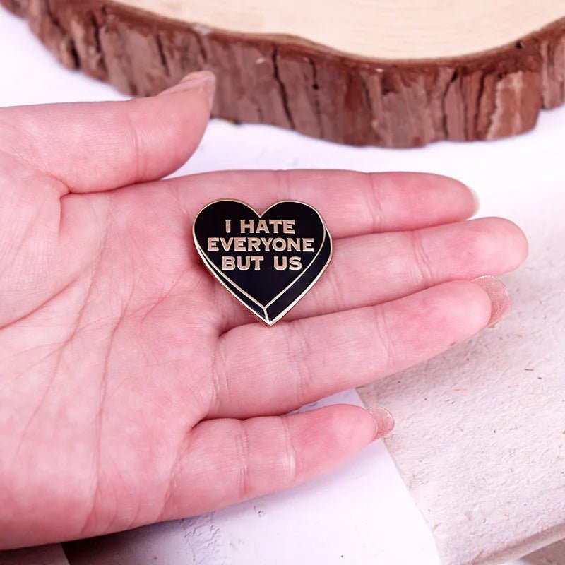 A hand holds a black heart enamel pin with gold edges and gold writing that says I HATE EVERYONE BUT US.