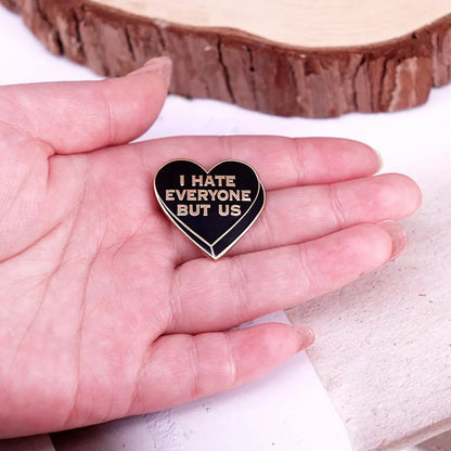 A hand holds a black heart enamel pin with gold edges and gold writing that says I HATE EVERYONE BUT US.