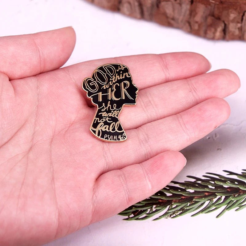 A hand holds a Kiss Me Later black silhouette enamel pin with gold text, shaped like a womans profile and reading God Is Within Her She Will Not Fall—Psalm 46:5. Pine needles and wood can be seen in the background.