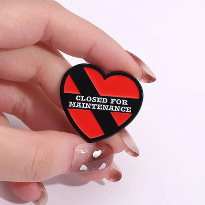 A hand with painted nails holds a heart enamel pin, which has a red background, a black diagonal stripe, and CLOSED FOR MAINTENANCE printed in white across the front.