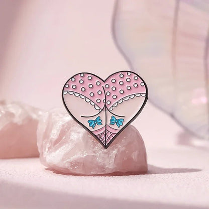 A heart-shaped enamel pin featuring a stylized closed-eye face, pink and white polka dots, and mermaid blue accents, rests on a pale pink crystal against a soft pastel background.