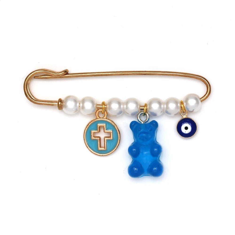 The Kiss Me Later gold safety pin, adorned with white beads, features three hanging charms—a blue and gold cross, blue gummy bear, and blue evil eye—making it a charming baby shower gift and unique evil eye pin.