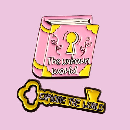 A pink illustrated book badge with a keyhole and Explore The World sits above a yellow Explore the world key enamel pin all set against a light purple background.