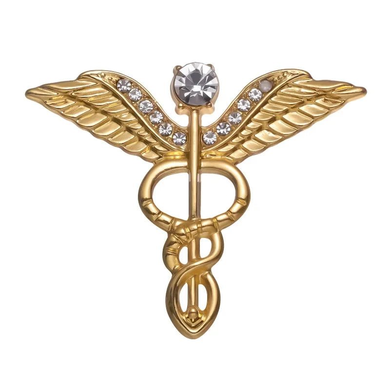 Gold medical symbol pin features two wings, a central rod with a snake, small white gemstones, and a large top gemstone—an elegant feminist gift.