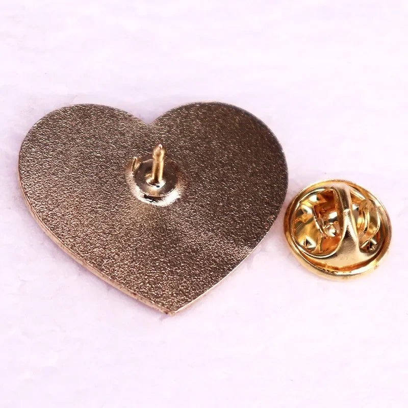A metallic pink love heart enamel pin shown from the back on a light surface, reveals its secure butterfly clutch attachment.