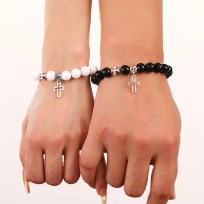 Two hands wear matching Kiss Me Later crystal bead bracelets, one with white and one with black beads, each featuring a small silver cross charm. A plain white background highlights these elegant Christian stone bracelets.