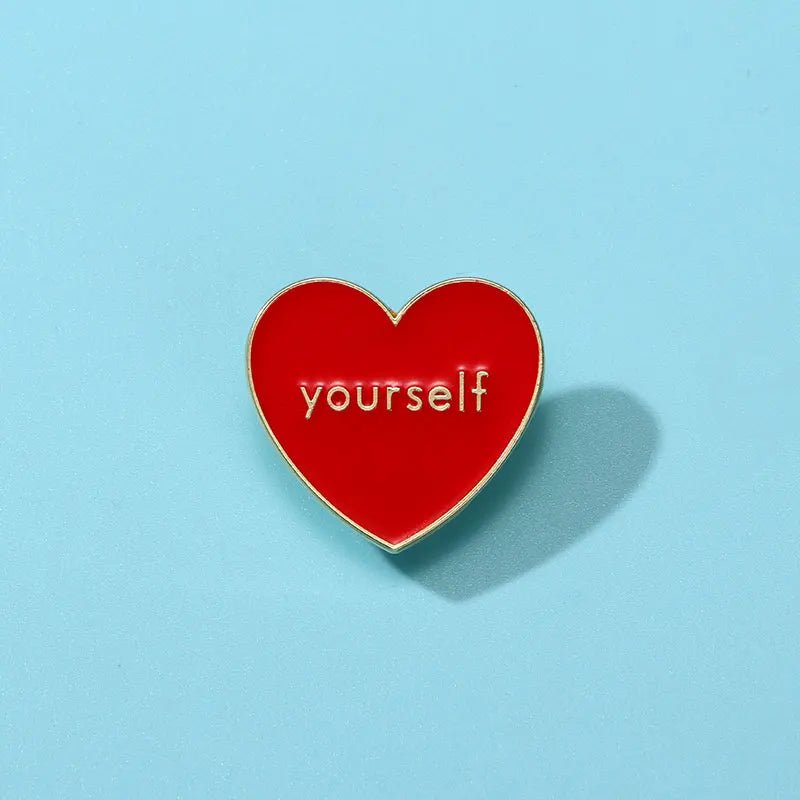 A Kiss Me Later red and gold enamel heart pin featuring yourself in white lowercase letters, displayed on a light blue background with a soft shadow on the right.