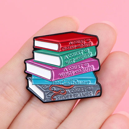 A hand holds an enamel pin shaped like a stack of five books, each titled from the “A Court of Thorns and Roses” series, perfect for book lovers, against a pink background.