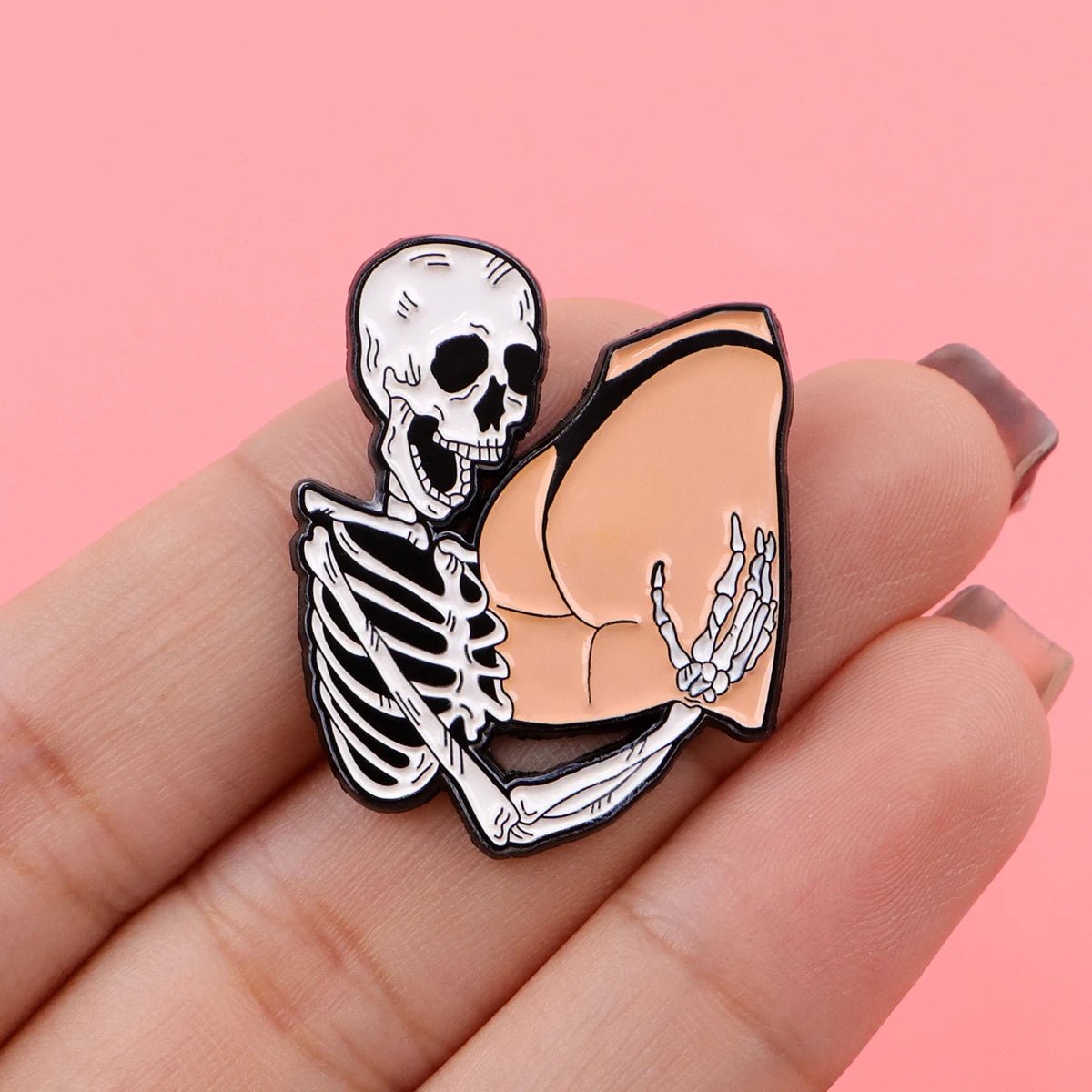 A hand displays a Funny Skeleton enamel pin, featuring a grinning skeleton hugging cartoon-style human buttocks, set against a playful pink background.