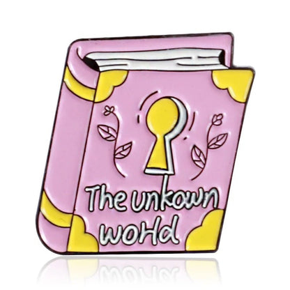 A pink enamel pin shaped like a book with yellow corners, a keyhole, flowers, and The unknown world text—a perfect badge for those ready to explore.
