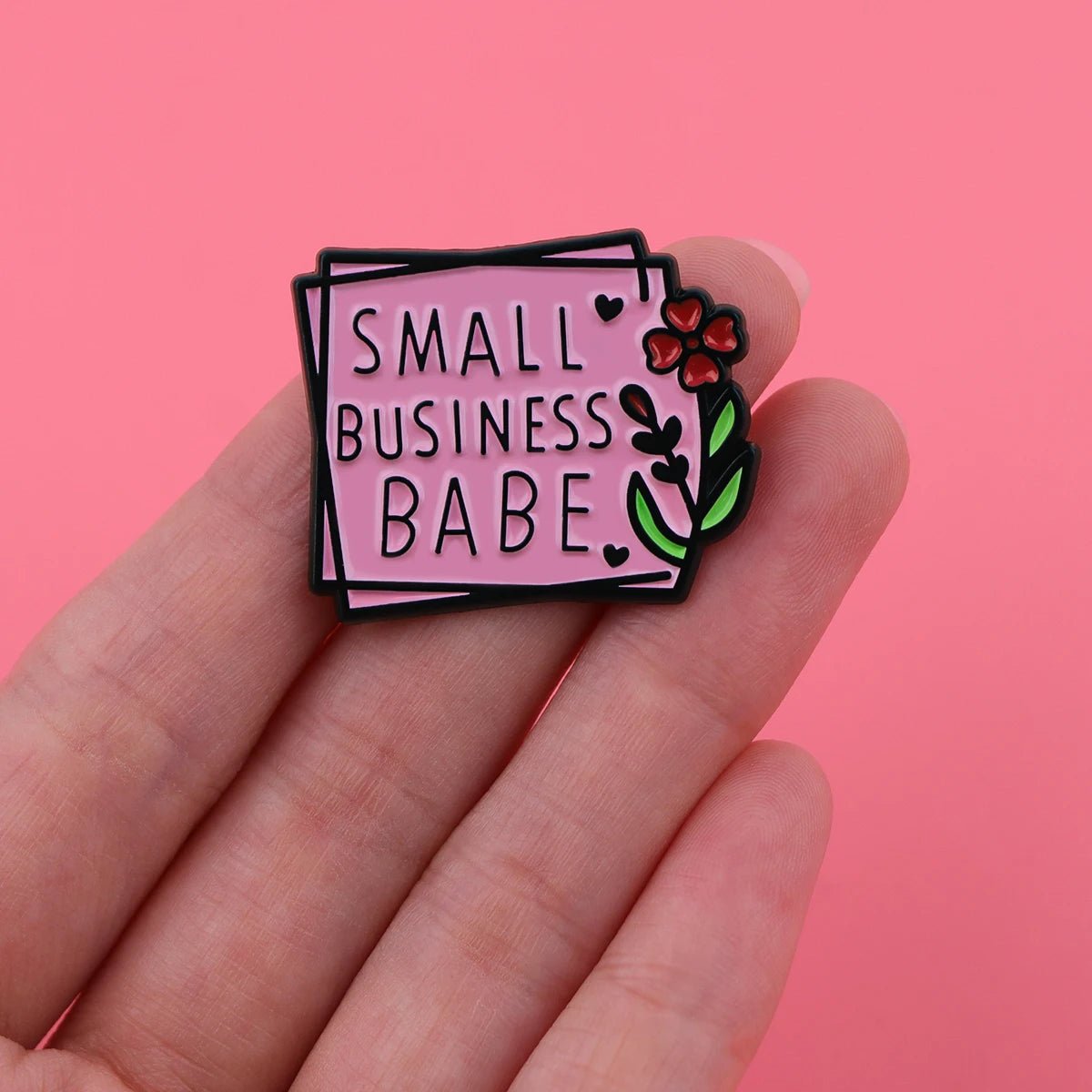 A hand holds a pink enamel pin with SMALL BUSINESS BABE in black letters, plus a red flower and green leaves—ideal for self-empowerment or sharing positive affirmations—set against a pink background.