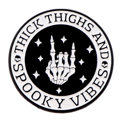 The Kiss Me Later enamel pin is a round black-and-white design with a skeleton hand rock-on gesture, stars, and the words Thick Thighs and Spooky Vibes circling the edge for an extra dose of spooky style.