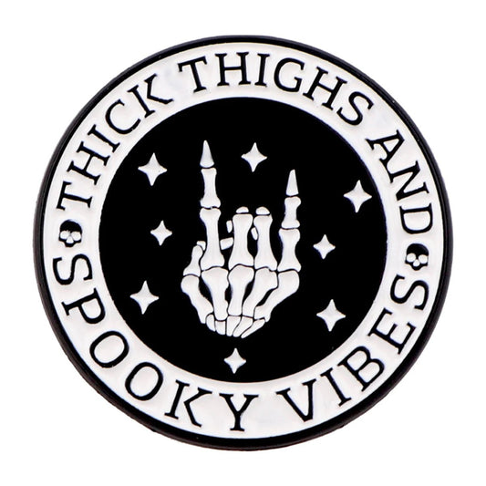 The Kiss Me Later enamel pin is a round black-and-white design with a skeleton hand rock-on gesture, stars, and the words Thick Thighs and Spooky Vibes circling the edge for an extra dose of spooky style.