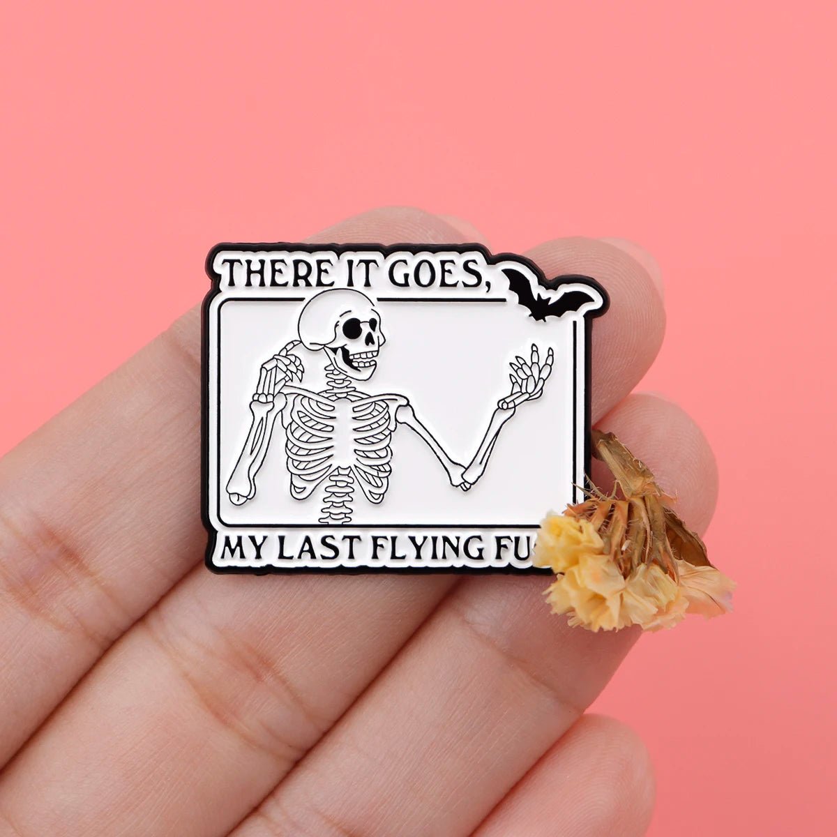 A hand holds a Kiss Me Later skeleton hand enamel pin featuring a waving skeleton and There it goes, my last flying fu. The pink background and dried yellow flowers enhance the spooky vibes.