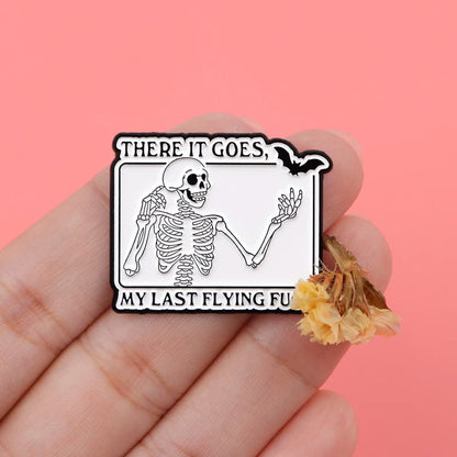 A hand holds a Kiss Me Later skeleton hand enamel pin featuring a waving skeleton and There it goes, my last flying fu. The pink background and dried yellow flowers enhance the spooky vibes.