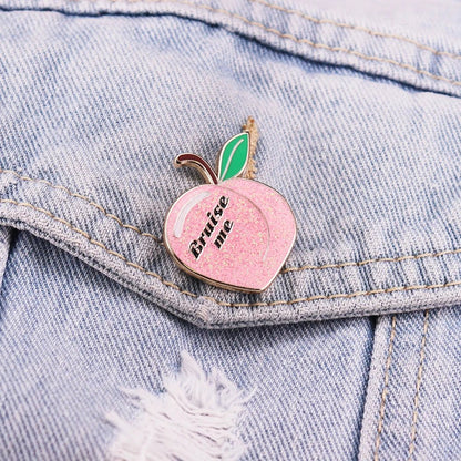 Gittery peach enamel pin, shaped like a peach with green leaves and Bruise Me text, is pinned to a light blue distressed denim jacket.
