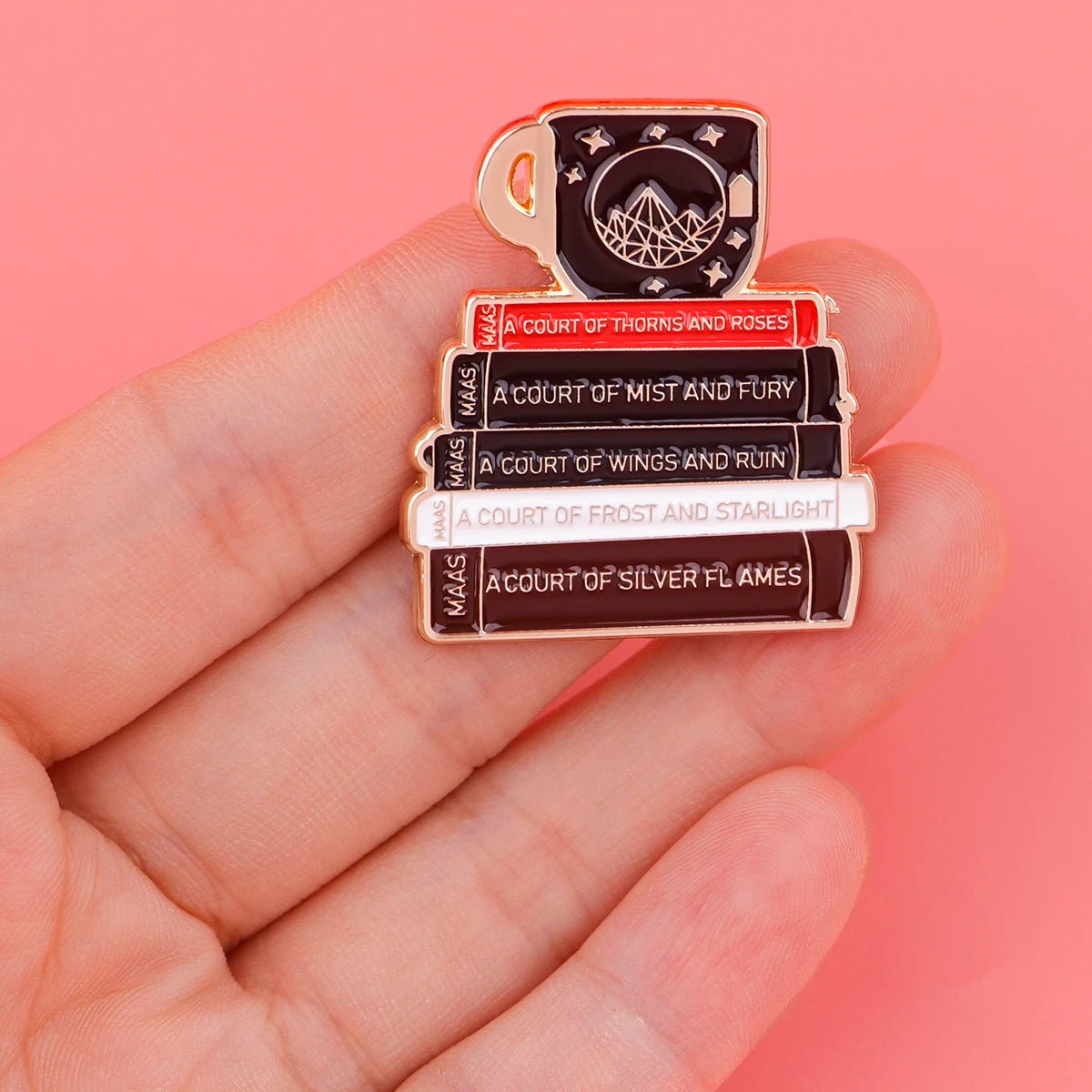 A hand holds an enamel pin shaped like a book stack topped with a mug, featuring titles from Sarah J. Maass A Court of Thorns and Roses series, ideal for any Booktrovert. Displayed on a pink background.